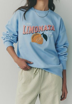 Femme portant un sweat-shirt bleu clair avec le texte « LIMONATA » et un motif citron, associé à un pantalon texturé clair.