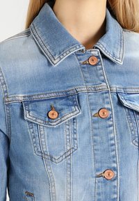 Jasnoniebieska kurtka dżinsowa z klasycznym kołnierzem, dwoma kieszeniami na piersiach i miedzianymi guzikami. Charakteryzuje się widocznym przeszyciem i wyblakłą teksturą.