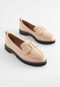 Light pink leather loafers with a contrasting black sole, featuring a decorative bow and gold-tone accent on the front. Smooth texture.