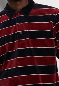 Close-up van een persoon die een fluwelen poloshirt draagt met horizontale kastanjebruine en zwarte strepen, een marineblauwe kraag en drie zwarte knopen.