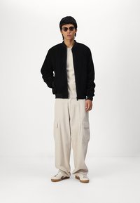 Jachetă bomber neagră din fleece cu fermoar în față, un hanorac crem dedesubt, pantaloni cargo bej și adidași albi cu talpă maro.