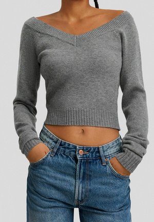 Femme portant un pull court gris à épaules dénudées avec des manches longues et un jean bleu taille haute, les mains dans les poches, debout devant un fond uni.