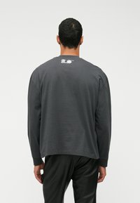 Γκρίζο μακρυμάνικο μπλουζάκι σε χαλαρή γραμμή με ραμμένα μανίκια, διαθέτει λευκό γραφικό λογότυπο στο πάνω μέρος της πλάτης. Απαλή βαμβακερή υφή.