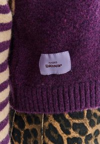Fialový pletený svetr s žebrovanými manžetami a texturovaným povrchem, s malou bílou štítkem s nápisem "NAOKO Knitwear." Viditelný pruhovaný vzor na rukávu.