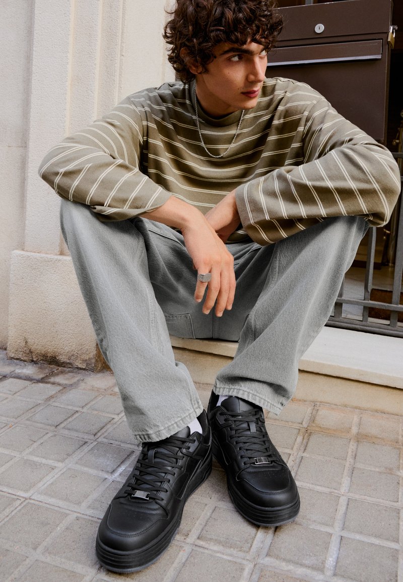 Jeune homme aux cheveux bouclés portant une chemise rayée, un pantalon gris clair et des baskets noires assis sur une marche à l'extérieur.