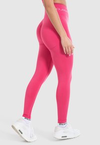 Leggings rosa brillante con vita alta, realizzati in materiale elasticizzato, con una texture liscia e una fascia con logo, abbinati a sneaker bianche.