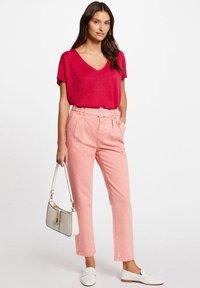 Rode V-hals top, lichtroze high-waisted broek met riem en witte instappers. Accessoire is een kleine witte handtas met contrasterende rand.