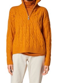 Maglione arancione a maglia grossa con colletto alto e zip, indossato sopra un dolcevita abbinato. I polsini e l'orlo a coste aggiungono texture.