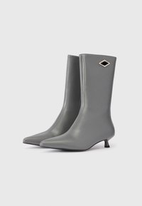 Pāris elegantu pelēku potīšu zābaku ar norādītām pirkstiem, maziem kaķa papēžiem un dimanta formas melnu un sudraba emblēmu pie augšas.