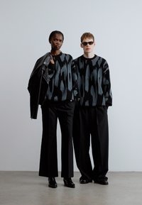 Dva modela nosita črne in sive puloverje z vzorci. En model ima usnjeno jakno; oba nosita črne hlače z širokimi hlačnicami in čevlje.