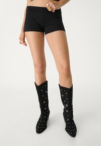 Zwarte slimfit shorts met een gladde textuur, gecombineerd met zwarte kniehoge laarzen met metalen ogen en een puntige neus.