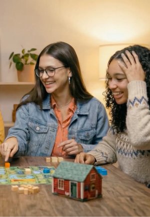 Two women smiling and playing a tabletop board game with colorful pieces and a small model house in a cozy room.