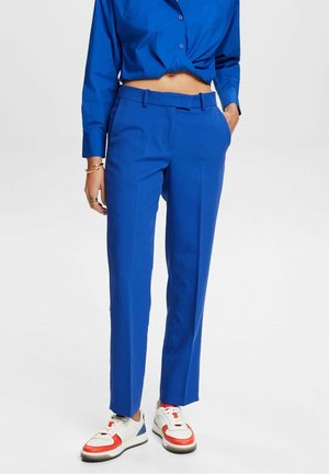 Persona vistiendo pantalones de tiro alto azul brillante y camisa a juego, con zapatillas blancas que tienen detalles en rojo y azul, de pie con una mano en el bolsillo.