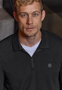 Svart zip-up pullover med texturerat tyg, med en logotypakcent på bröstet, lager på en vit t-shirt. Nära bild.