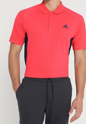 Mężczyzna w koralowo-różowej koszulce polo Adidas i ciemnych spodniach ze ściągaczem, widoczny tors, ręce swobodnie opuszczone wzdłuż ciała.