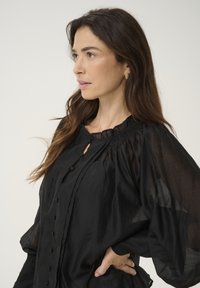 Blusa nera con scollatura arricciata, dettagli sui bottoni e maniche lunghe voluminose. Realizzata in tessuto leggero e trasparente con una texture morbida.