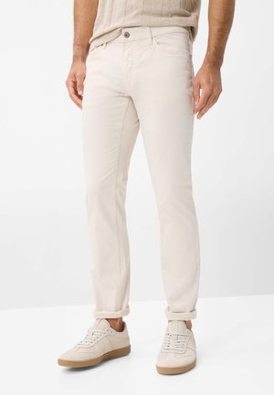 Mann trägt beige, slim-fit hochgekrempelte Hose mit passenden beigen Sneakern und einem hellbraunen, gerippten Pullover, steht auf weißem Boden.