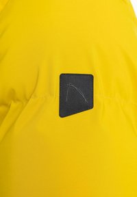 Gul udendørs jakke stof med en glat tekstur; har en sort firkantet applikation med et logo i det øverste højre hjørne.