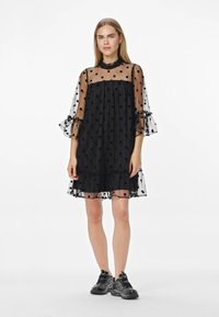 Crna haljina na točkice s prozirnim slojem, naboranim rukavima i proširenim krojem. Nošena s crnim sportskim tenisicama.