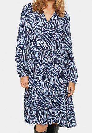 Kobieta ubrana w sukienkę z długim rękawem do kolan o abstrakcyjnym wzorze zebry w kolorach niebieskim, czarnym i różowym, zestawioną z czarnymi kozaczkami sięgającymi do kolan.