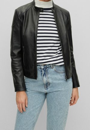 Person iført en sort læderjakke over en hvid og sortstribet skjorte, kombineret med lyseblå højhøjede jeans mod en ensfarvet baggrund.