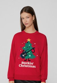 Junge Frau, die einen roten Pullover mit einem Comic-Weihnachtsbaum trägt, während sie eine Gitarre hält und den Text "Rockin' Christmas" auf der Vorderseite hat.