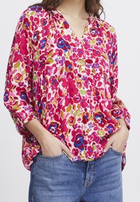 Kvinna iklädd en lös, knäppt blommig blus i röd, blå, grön och lila, tillsammans med blå jeans, med händerna i fickorna.