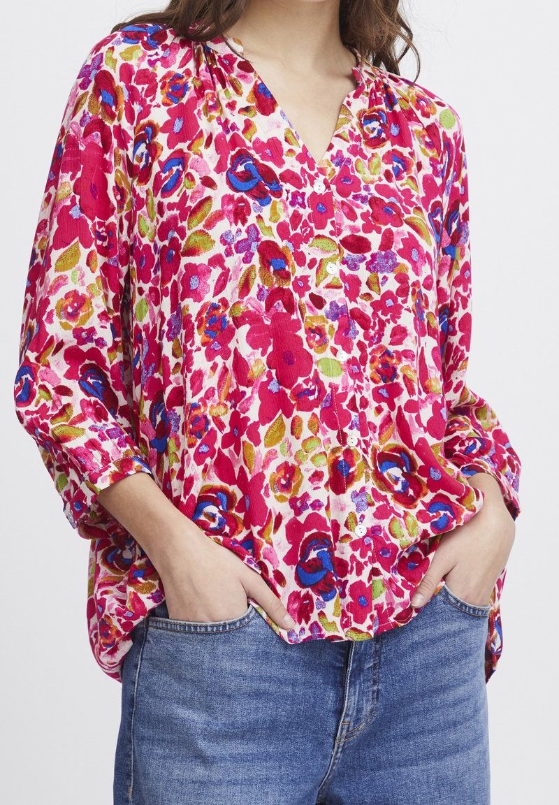 Kvinna iklädd en lös, knäppt blommig blus i röd, blå, grön och lila, tillsammans med blå jeans, med händerna i fickorna.