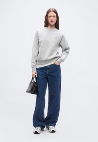 Femme portant un sweat-shirt gris clair, un jean large bleu, des baskets blanches, tenant un petit sac à main noir, debout devant un fond uni.