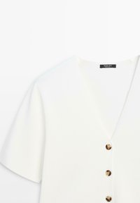 Cardigan blanc à manches courtes avec col en V, trois boutons marron et une étiquette au niveau de l'encolure sur un fond uni.