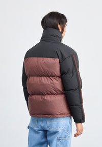 Chaqueta acolchada negra y burdeos con un cuello alto, que presenta un diseño de compartimentos, patrones texturizados sutiles en las secciones burdeos y detalles en rojo.