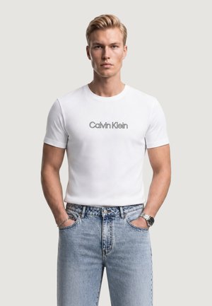 Blondyn w białej koszulce Calvin Klein i jasnoniebieskich dżinsach, z rękami w kieszeniach, stojący na jednolitym tle.