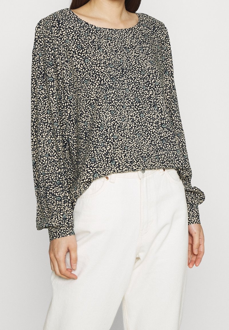 Langärmlige Bluse mit rundem Ausschnitt, marineblauem Hintergrund und weißem Blumenmuster. Der Stoff wirkt weich und hat eine lockere Passform. Trägt man mit hellfarbigen Hosen.