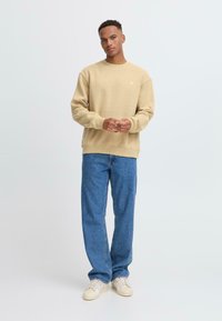 Beige sweatshirt with ribbed cuffs, crew neck, and small logo. Paired with light blue straight-leg jeans and beige sneakers. Plain background.