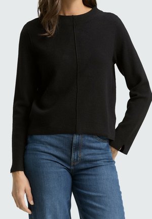 Femme portant un pull noir uni à manches longues avec une couture centrale, associé à un jean bleu, debout avec une main dans la poche.