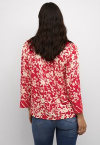 Blusa rossa floreale con maniche lunghe e vestibilità rilassata. Presenta fiori color crema e un dettaglio a righe nei polsini. Abbinata a jeans blu.