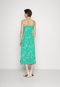 Vestido midi verde com padrão floral branco, alças ajustáveis finas, corpete ajustado e saia evasé. Usado com sandálias de salto bloco em tom pálido.