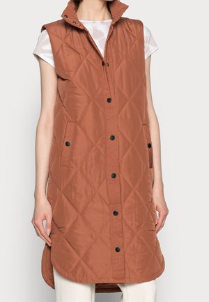 Bezrekawnik o rdzawej brązowej pikowanej tkaninie z wysokim kołnierzem, czarne zatrzaski, boczne kieszenie, noszony na białej bluzce z krótkim rękawem i jasnych spodniach.