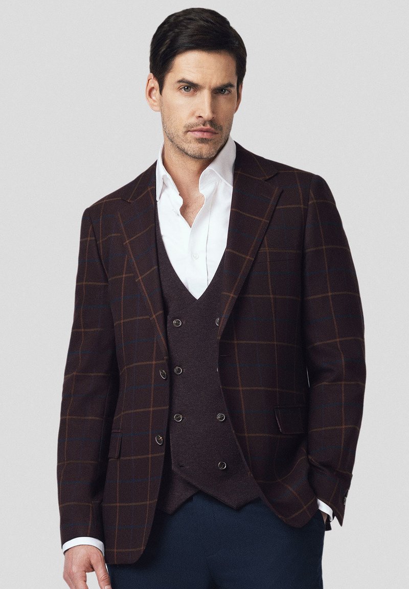 Brązowa marynarka w kratę z kontrastującymi niebieskimi i pomarańczowymi liniami, noszona na ciemnej kamizelce i białej koszuli, z dwoma guzikami i wyłogami w kształcie szpitu.