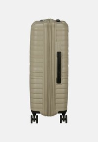 Valigia rigida di colore beige chiaro con scanalature orizzontali, doppia zip, manico laterale e quattro ruote nere per una facile manovrabilità.