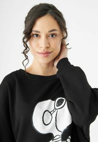 Sweatshirt noir avec un grand motif Snoopy blanc. Texture douce, coupe décontractée et poignets côtelés mettent en valeur le design.