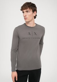 Langermet T-skjorte med rund hals i grått stoff, med en stor svart "Armani Exchange" logo på forsiden. Glatt tekstur, tettsittende design.
