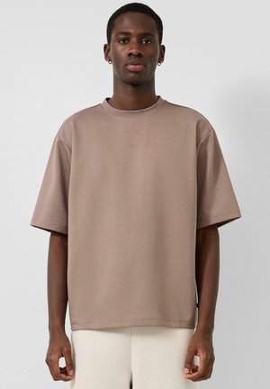 Jeune homme noir portant un t-shirt ample à manches courtes couleur taupe et un pantalon crème, debout contre un mur blanc uni.
