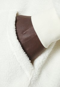 Weißer Fleece-Stoff mit weicher Textur; eine dunkelbraune Hand ragt aus einer Tasche, trägt eine cremefarbene, gerippte Manschette.