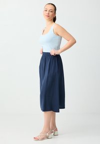 Jeune femme debout, les mains sur les hanches, portant un débardeur bleu clair, une jupe midi bleu marine et des sandales blanches à talons carrés avec brides.