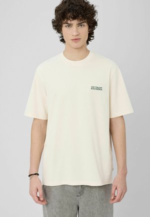 T-shirt en coton couleur crème à manches courtes, présentant un petit texte vert indiquant "EAT ITALIAN KISS FRENCH." Coupe décontractée.