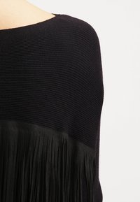 Svart ribbat stickat tyg med ett horisontellt band av långa svarta fransar som detaljerar under axelområdet.