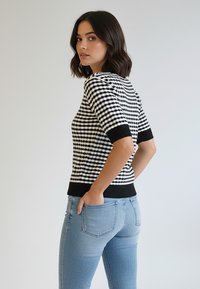 Sweter z dzianiny w czarno-białą kratę z krótkimi rękawami i czarnym prążkowanym dołem, zestawiony z jasnoniebieskimi dżinsami.