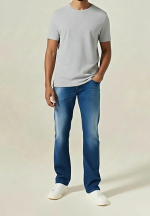 Mann trägt hellgraues T-Shirt mit kurzen Ärmeln, blaue Jeans mit ausgewaschenen Stellen, weiße Turnschuhe, steht mit einer Hand in der Tasche.