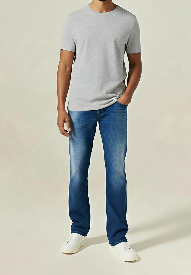 Mann trägt hellgraues T-Shirt mit kurzen Ärmeln, blaue Jeans mit ausgewaschenen Stellen, weiße Turnschuhe, steht mit einer Hand in der Tasche.